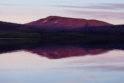 Reflection of distant sunset boreal forest taiga mountain mirrored on calm water surface of Little Salmon Lake, Yukon Territory, Canada