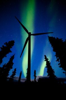 Wind turbine supplying renewable electric energy from wind at night surrounded by the silhouettes of tall trees against a starry sky with green aurora borealis northern lights