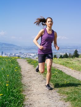 Photo of a young woman jogging and exercising on a country path.  Lake and city in the distance. Slight motion blur visible.