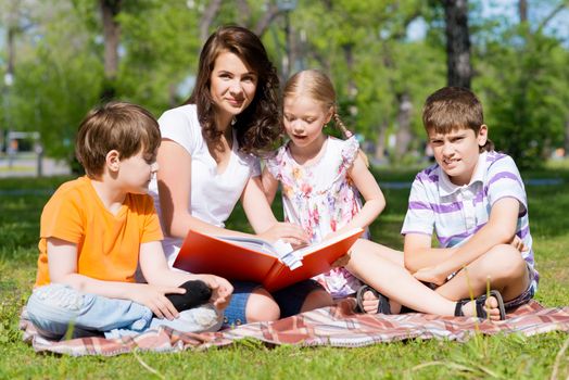 children and teacher reading book together in the summer park