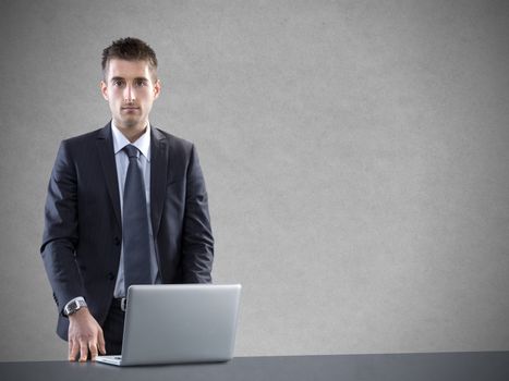 Young businessman with laptop leaning on desk against gray background.