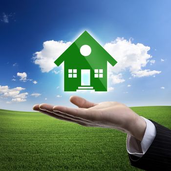 Businessman hand showing a green house icon on meadow and blue sky background.