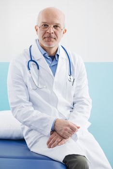 Friendly confident doctor sitting on a medical examination bed.