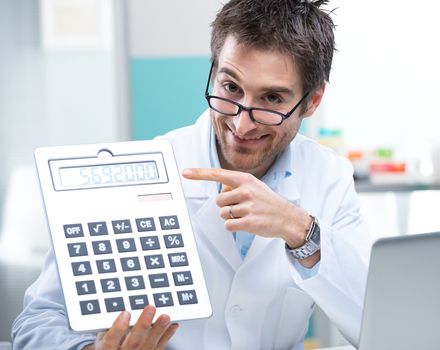 Young friendly doctor pointing at a big calculator.