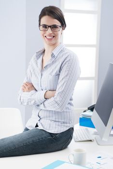 Attractive young businesswoman sitting on desk and smiling confidently at camera with arms crossed.