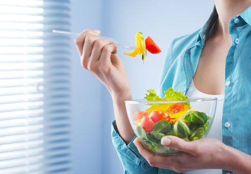 Woman holding a bowl and eating salad in front of a window.