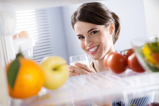 Attractive woman smiling and holding a glass of milk in front of refrigerator.