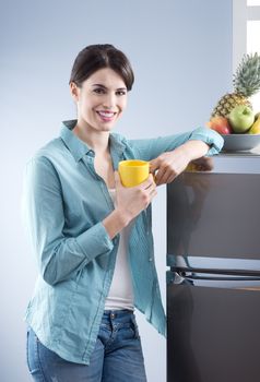 Young cheerful woman holding a yellow mug and leaning against refrigerator.