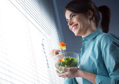 Woman eating salad in front of a window and smiling.