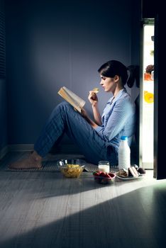 Sleepless woman sitting on the kitchen floor reading a book and eating.