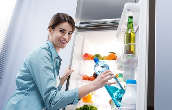 Young woman taking a water bottle from refrigerator and smiling at camera.