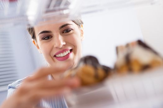 Young woman smiling and taking pastry with chocolate topping from fridge.