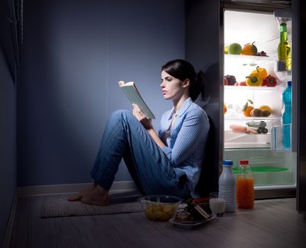 Sleepless woman sitting on the kitchen floor reading a book and eating.