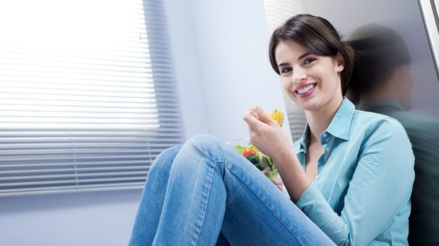 Young woman sitting in front of the fridge eating salad.