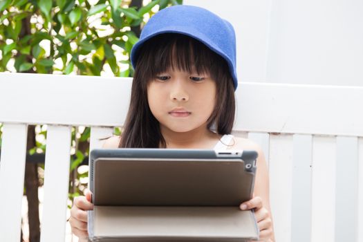 Girl playing with tablet on bench. girl with long black hair wearing blue hat.