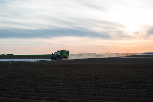 Modern tractor with trailer driving along agricultural field and raising dust due to dry weather conditions in early spring with great dramatic sunset