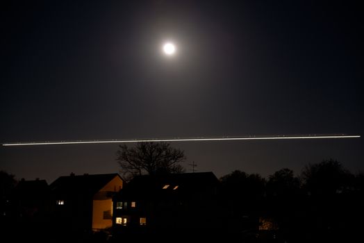 Bright moon shining over houses and trees with traces of a landing airplane