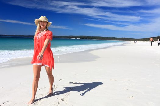 Happy woman walking along a beautiful white sandy beach in sunny Australia.  She is barefoot, wearing a sun hat, white shorts and a floaty top