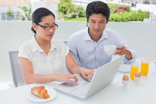 Smiling couple having breakfast together using laptop outside on a balcony