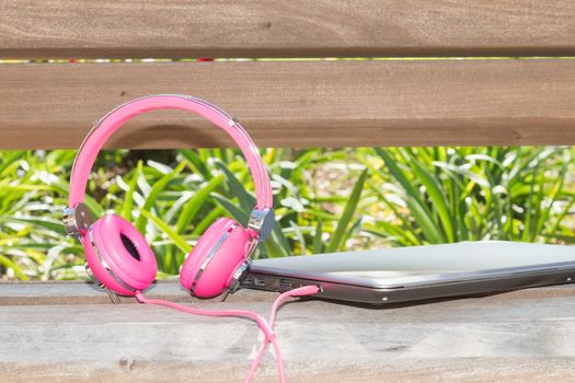 Vividl pink headphones and laptop on the wooden bench in park