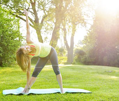 Female fitness instructor doing yoga extended triangle pose outdoors in morning sunshine