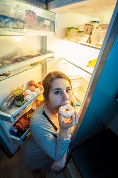 Portrait of young woman in pajamas eating donut next to refrigerator