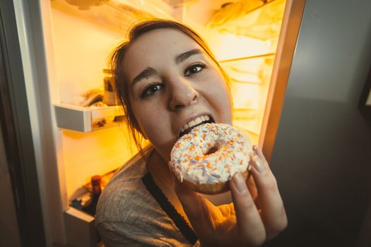 Wide angle portrait of hungry woman eating donut on kitchen at night