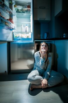 Photo of woman in pajamas eating on floor next to fridge at night