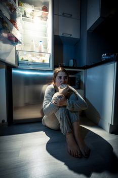 Photo of young woman eating next to refrigerator at night