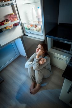Hungry woman sitting on floor and looking at fridge at night