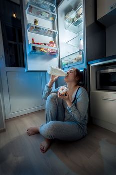 Portrait of young woman sitting on floor at kitchen and having late dinner