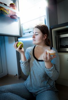 Portrait of woman holding apple and donut against fridge at night
