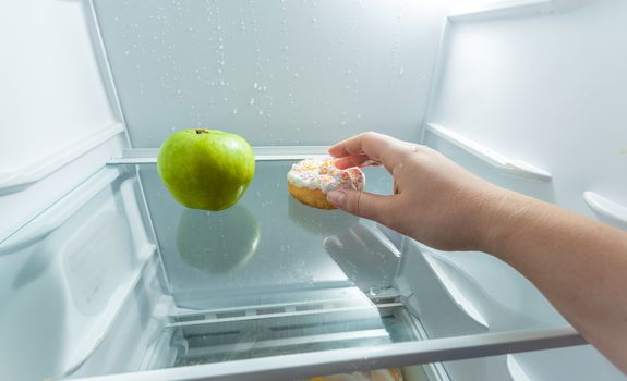 Closeup photo of hand choosing donut instead of apple lying in fridge