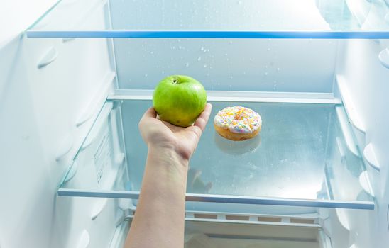 Closeup photo of women hand holding apple instead of donut in refrigerator