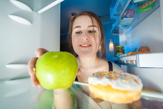 Portrait from inside the fridge of woman taking green apple
