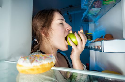 Closeup portrait from inside of fridge of young woman eating green apple