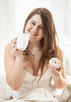 Closeup portrait of smiling woman holding cup of coffee and donut in bed