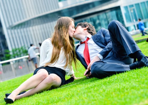 Successful lovers laying down on green grass near a modern glass office building, listening music and using laptop