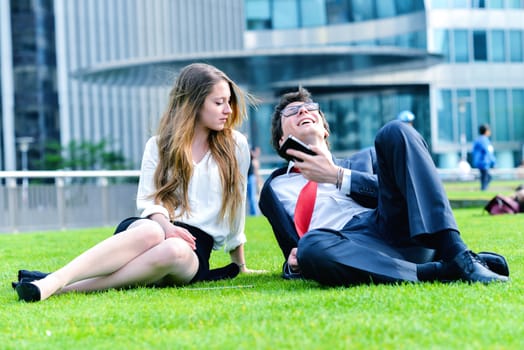 Successful Junior executives dynamics laying down on green grass near a modern glass office building, listening music on smartphone