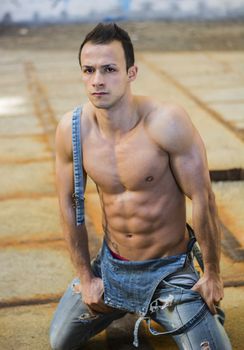 Muscular shirtless young man with jeans overalls kneeling, looking at camera