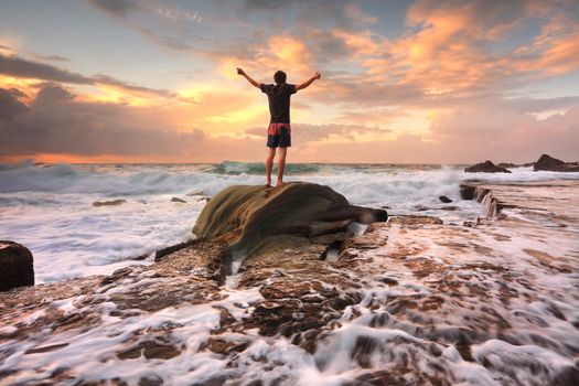 Teen boy stands on a rock among turbulent ocean seas and fast flowing water at sunrise.  Worship, praise, zest, adenture, solitude, finding peace among lifes turbulent times.  Overcoming adversity.  Motion in water