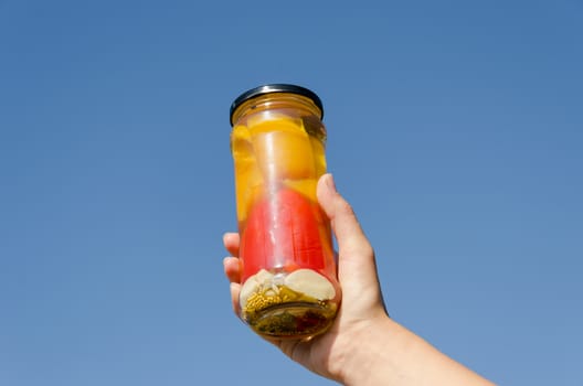 female hand hold marinated pepper in glass jar on blue sky background