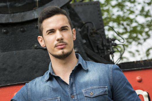 Handsome young man in denim shirt in front of old train, looking at camera