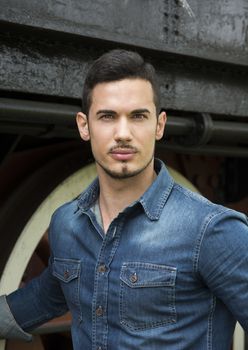 Handsome young man in denim shirt in front of old train, looking at camera