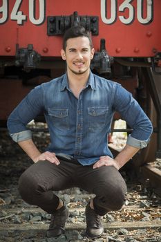 Handsome young man in denim shirt sitting in front of old train, looking at camera