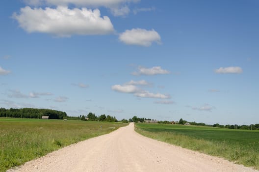 gravel country road continues into distance along the green fields on blue sky background