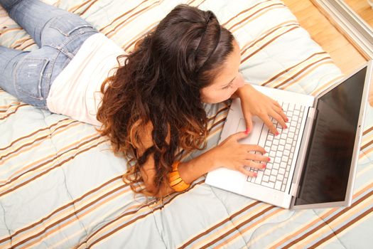 A young girl laying on the bed and surfing on the Internet with a Laptop.  