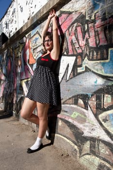 A vintage style dressed girl leaning at a graffiti wall and enjoying the sunlight.