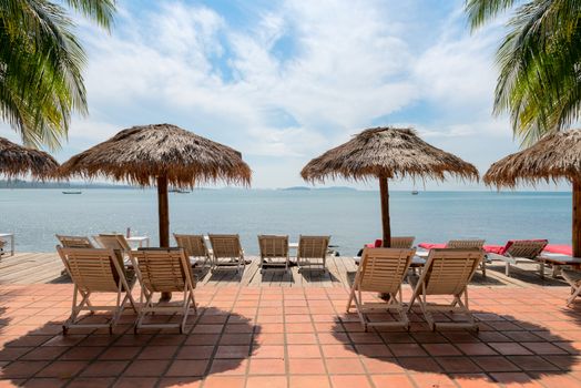 Vacation Concept. Palms and relaxing sunbeds on tile and wooden desk near the sea under blue sky.