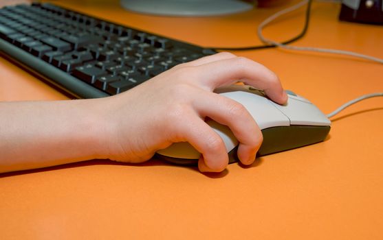 Children's right hand on the mouse on the background of computer keyboard on the orange table